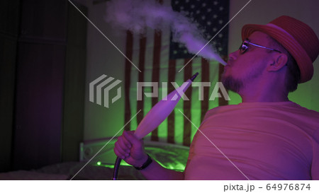 Hookah Smoker Exhaling Puffs Of Smoke In Home Interior With Neon Lights 64976874