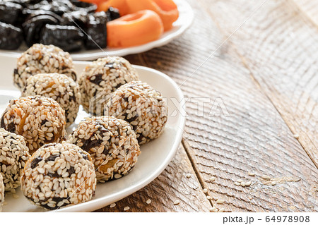 Healthy handmade energy balls made from pieces of dried apricots, prunes and sesame seeds on white plate and plate of whole dried apricots and prunes on wooden table. Copy space, selective focus.  64978908