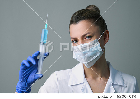 A female medical worker in a white robe and mask holds a syringe. 65004084