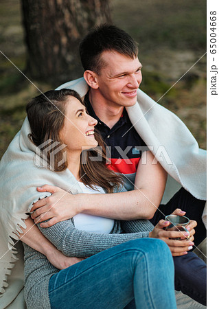 A couple in love sits on the riverbank at sunset. They cuddle, roll into a plaid and drink tea from a metal hiking mug. 65004668