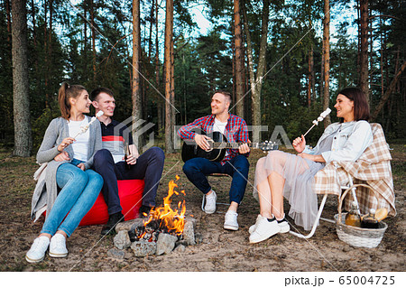 A group of friends relax in a forest camp. Men and women prepare a marshmallow on a bonfire. A party in nature. 65004725