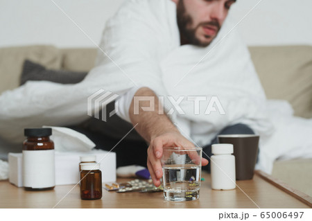 Sick young man wrapped into blanket sitting on couch and taking glass of water Sick young man wrapped into blanket sitting on couch and taking glass of water 65006497