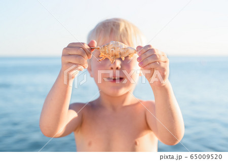 Cute blond boy holding a shell on sea background. 65009520