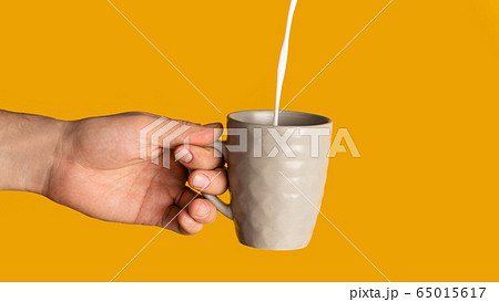 Unrecognizable young man holding cup of hot drink with milk pouring into it, orange background. Panorama Unrecognizable young man holding cup of hot drink with milk pouring into it, orange background. Panorama 65015617