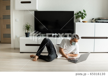 Man in medical mask is lying on a floor in his living room and working from home with a laptop because of coronavirus. Remote work during pandemic. Stay home during COVID-19 quarantine. Freelancer. 65016130