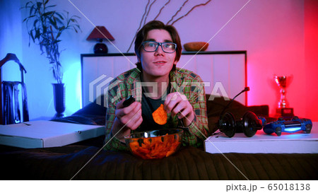 Young interested man in glasses lying on the bed watching TV and eating chips 65018138