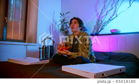 Young interested man sitting on the bed watching TV and eating chips 65018180