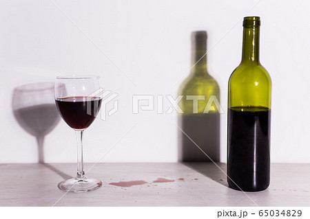 Bottle with wine and glass, red puddle of wine on the table. Cleaning after party concept. Bottle with wine and glass, red puddle of wine on the table. Cleaning after party concept. 65034829
