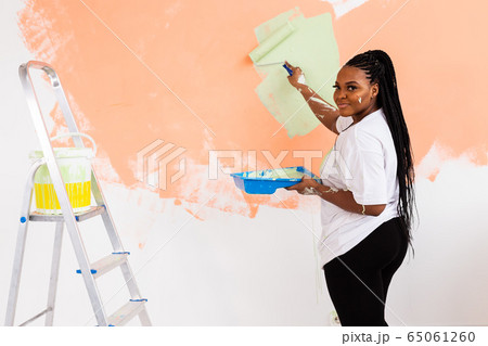 Beautiful african american girl painting the wall with paint roller. Portrait of a young beautiful woman painting wall in her new apartment. Redecoration and renovation concept. Beautiful african american girl painting the wall with paint roller. Portrait of a young beautiful woman painting wall in her new apartment. Redecoration and renovation concept. 65061260