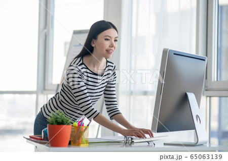 Dark-haired woman in a striped shirt standing near the table and looking at the computer Dark-haired woman in a striped shirt standing near the table and looking at the computer 65063139