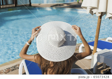 Beautiful woman sunbathing on a beach at tropical travel resort, enjoying summer holidays. Girl holding a hat with her hands on sun lounger near the pool 65073428