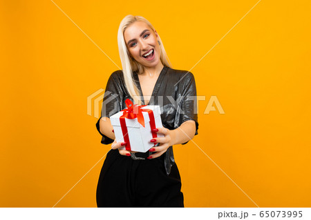 blond girl holds out a gift box with a red ribbon on a yellow studio background blond girl holds out a gift box with a red ribbon on a yellow studio background 65073995