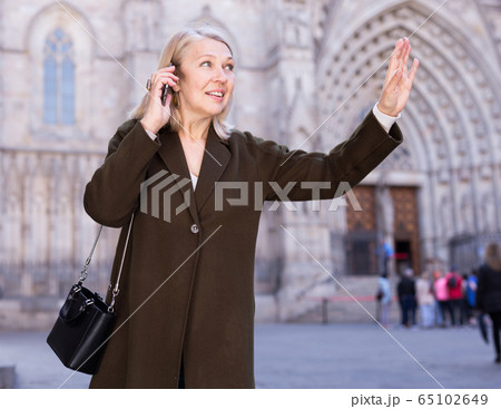 Happy woman speaks on the phone and greets someone on the street with her hand 65102649