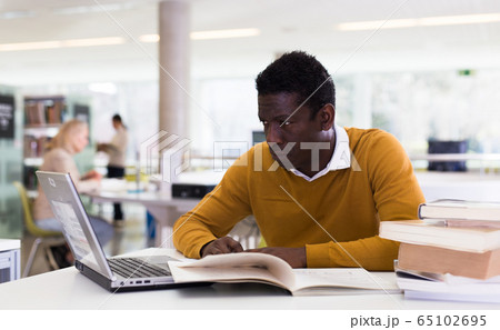 Efficient man preparing for exam in public library, reading book and using laptop for searching information Efficient man preparing for exam in public library, reading book and using laptop for searching information 65102695