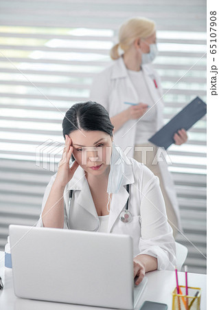 Brunette woman behind a laptop, blond-haired in mask. Brunette woman behind a laptop, blond-haired in mask. 65107908