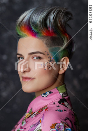 Portrait of a young beautiful girl in a pink shirt on a gray background with dyed hair. Short haircut pixie or bob. Rainbow coloring and sequins on the face. 65108891