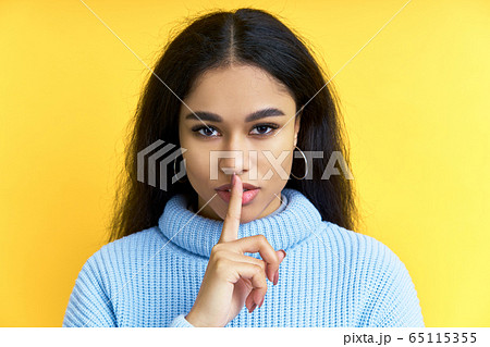 Pretty black woman keeps finger on lips, making hush gesture on yellow background 65115355