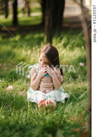 Cute little girl sits on the grass barefoot and hug teddy bear. Kid spend time in the park, outside. Beautiful little girl model Cute little girl sits on the grass barefoot and hug teddy bear. Kid spend time in the park, outside. Beautiful little girl model 65120597