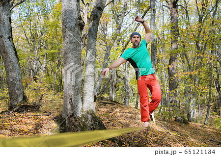 An aged bearded man in orange pants walks on a slack stretched between trees in an autumn forest. The concept of balance and sports lifestyle for people over 40 65121184