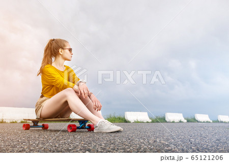 A young attractive girl in a yellow sweater shorts and sunglasses with a tattoo on her arm sits on a longboard behind a suburban asphalt pad against a cloudy sky. 65121206