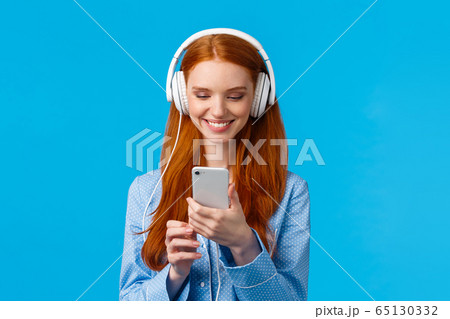 Technology addication, lifestyle and women concept. Cheerful good-looking redhead woman in nightwear, wearing big headphones listen music, smiling pick songs in smartphone app 65130332