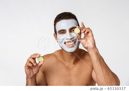 Photo of joyful african american man in facial cosmetic mask making fun with cucumber isolated over white background 65130337