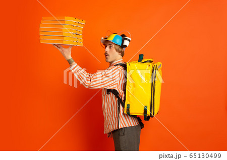 Contacless delivery service during quarantine. Man delivers food and shopping bags during insulation. Emotions of deliveryman isolated on orange background. Contacless delivery service during quarantine. Man delivers food and shopping bags during insulation. Emotions of deliveryman isolated on orange background. 65130499