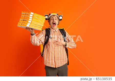 Contacless delivery service during quarantine. Man delivers food and shopping bags during insulation. Emotions of deliveryman isolated on orange background. Contacless delivery service during quarantine. Man delivers food and shopping bags during insulation. Emotions of deliveryman isolated on orange background. 65130535