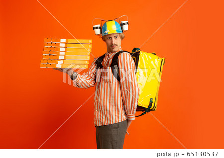 Contacless delivery service during quarantine. Man delivers food and shopping bags during insulation. Emotions of deliveryman isolated on orange background. 65130537