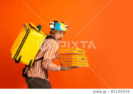 Contacless delivery service during quarantine. Man delivers food and shopping bags during insulation. Emotions of deliveryman isolated on orange background. Contacless delivery service during quarantine. Man delivers food and shopping bags during insulation. Emotions of deliveryman isolated on orange background. 65130543