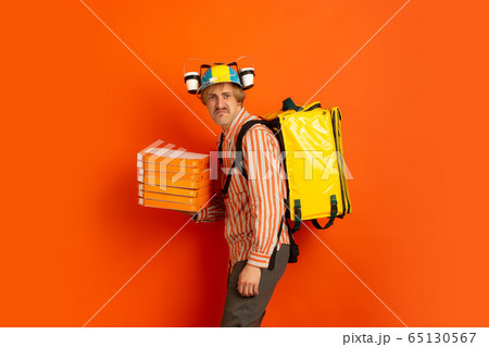 Contacless delivery service during quarantine. Man delivers food and shopping bags during insulation. Emotions of deliveryman isolated on orange background. Contacless delivery service during quarantine. Man delivers food and shopping bags during insulation. Emotions of deliveryman isolated on orange background. 65130567
