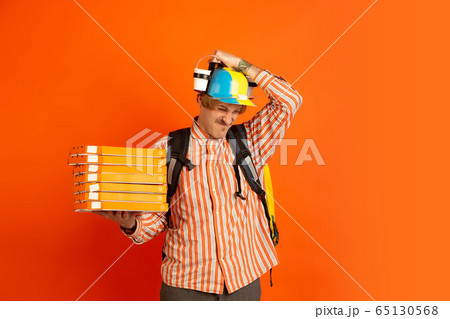 Contacless delivery service during quarantine. Man delivers food and shopping bags during insulation. Emotions of deliveryman isolated on orange background. 65130568