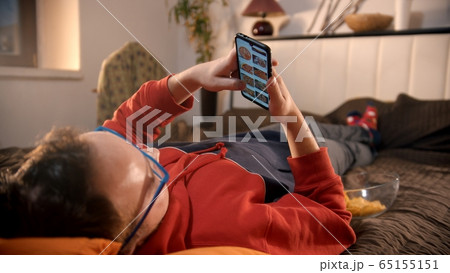 Young man lying on the bed and scrolling pictures of pizza on his phone and about to order it 65155151