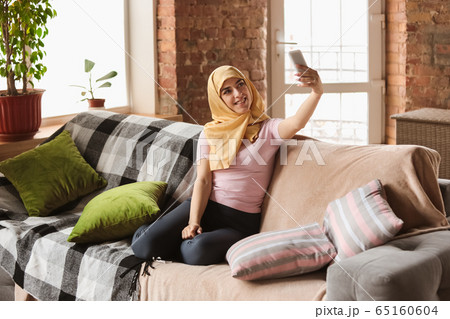 A pretty young muslim woman at home during quarantine and self-insulation, using smartphone for selfie or videocall, online lessons, shopping 65160604