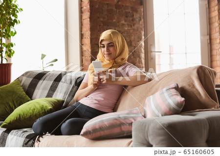 A pretty young muslim woman at home during quarantine and self-insulation, using smartphone for selfie or videocall, online lessons, shopping 65160605