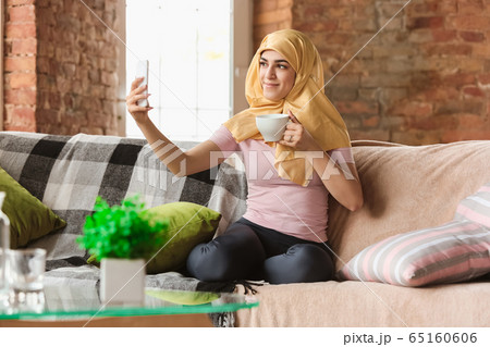 A pretty young muslim woman at home during quarantine and self-insulation, using smartphone for selfie or videocall, online lessons, shopping A pretty young muslim woman at home during quarantine and self-insulation, using smartphone for selfie or videocall, online lessons, shopping 65160606