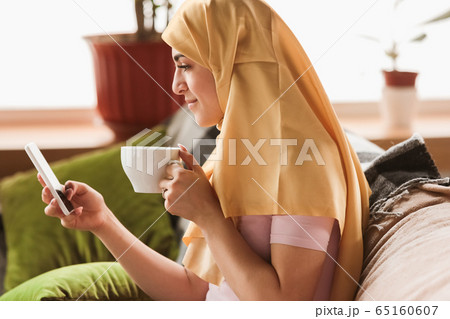 A pretty young muslim woman at home during quarantine and self-insulation, using smartphone for selfie or videocall, online lessons, shopping A pretty young muslim woman at home during quarantine and self-insulation, using smartphone for selfie or videocall, online lessons, shopping 65160607