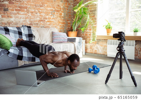 Young african-american man teaching at home online courses of fitness, aerobic, sporty lifestyle during quarantine, reording on camera, streaming Young african-american man teaching at home online courses of fitness, aerobic, sporty lifestyle during quarantine, reording on camera, streaming 65165059