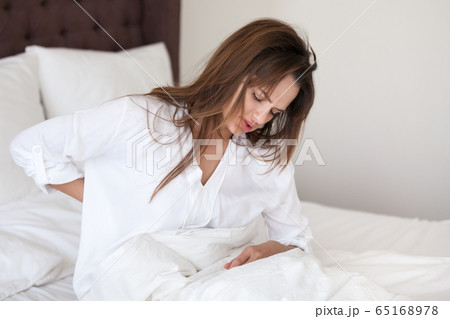 Upset woman stretching in bed feeling discomfort in aching back Upset woman stretching in bed feeling discomfort in aching back 65168978