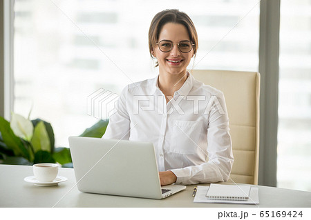 Smiling millennial businesswoman sitting at work desk with lapto Smiling millennial businesswoman sitting at work desk with lapto 65169424