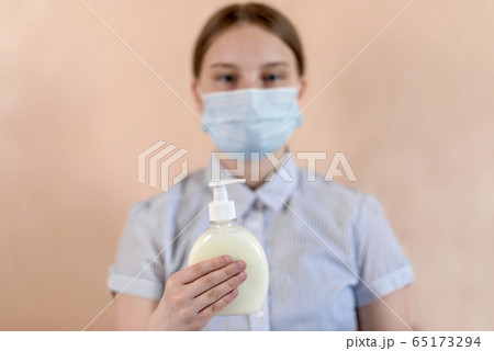 A teenager girl of 12-15 years old, in a medical mask covers her face, holds an antiseptic in her hand, liquid soap, care and help remind patients about safety, and washing hands. A teenager girl of 12-15 years old, in a medical mask covers her face, holds an antiseptic in her hand, liquid soap, care and help remind patients about safety, and washing hands. 65173294