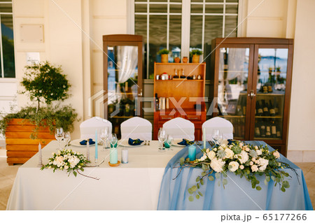 Wedding dinner table reception. White rectangular table of newlyweds with four chairs, white-blue tablecloth, floral arrangements with eucalyptus leaves, against background of wine cabinets 65177266