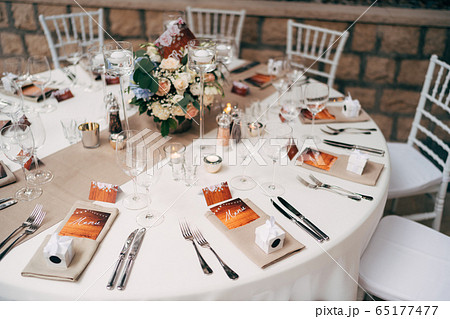 Wedding dinner table reception. Round table with white tablecloth with brown runner on table. Vienna nested in brown napkins, a bouquet of flowers is on the table. White Chiavari chairs with pillows. Wedding dinner table reception. Round table with white tablecloth with brown runner on table. Vienna nested in brown napkins, a bouquet of flowers is on the table. White Chiavari chairs with pillows. 65177477