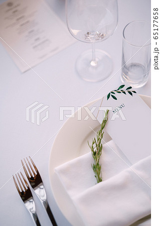 Wedding dinner table reception. White plate with cloth towel with paper menu and sprig of rosemary, against the background of white tablecloth 65177658