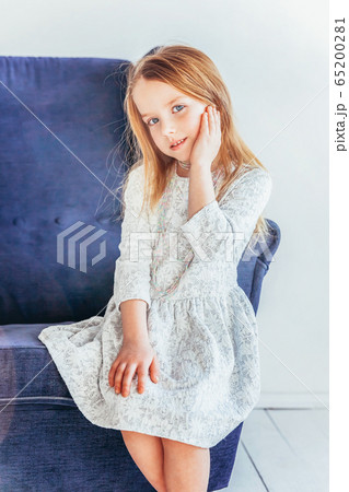 Stay Home Stay Safe. Sweet little girl in white dress sitting on modern cozy blue chair relaxing in white bright living room at home indoors. Childhood schoolchildren youth relax concept. 65200281