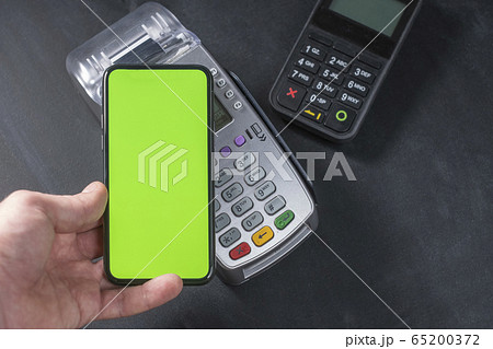 Top view of person using contactless payment with smartphone. Clear green mockup screen template of phone and pos terminal on dark wooden desk with side copy space 65200372
