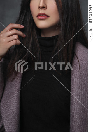 Beautiful young brunette girl in a black sweater and lilac coat on a gray background. Close-up, sensual lips and hands. 65201098
