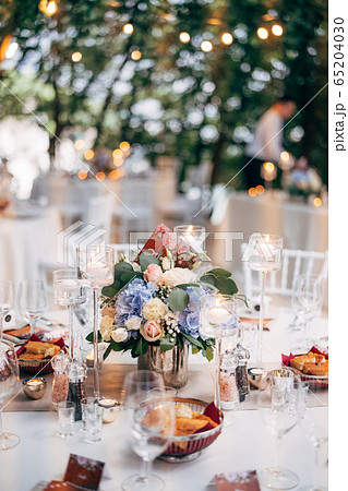 Wedding dinner table reception. Bouquet in a vase on a table of pink and white roses and blue hydrangea, with the number of the table on the plate. Burning candles in high glass candlesticks. Wedding dinner table reception. Bouquet in a vase on a table of pink and white roses and blue hydrangea, with the number of the table on the plate. Burning candles in high glass candlesticks. 65204030