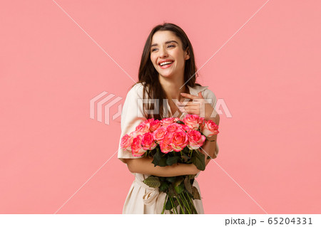 Happiness, romance and beauty concept. Cheerful gorgeous brunette girl in dress, holding roses and smiling flattered, touch chest laughing look away blushing, standing pink background enthusiastic 65204331