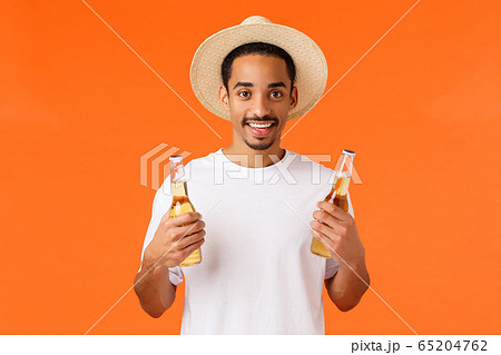 How bout drink. Handsome relaxed and happy smiling african-american man in hat, white t-shirt, holding two bottles of beer, enjoying weekends, relaxing with friends, having party, orange background 65204762
